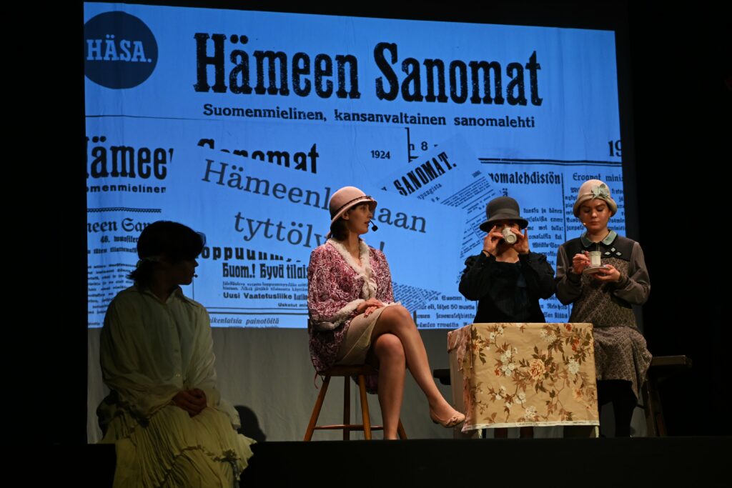 Näyttelijöitä lavalla pukeutuneina vanhoihin vaatteisiin. Taustalla näkyy Hämeen Sanomien uutisartikkeleita tyttölyseon perustamisesta.